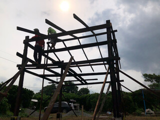 Fototapeta premium Silhouettes of carpenters on top of the wooden frame are joining girders and beams to construct a simple hut. Silhoettes