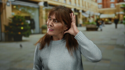 Fototapeta premium Senior hispanic woman on city street listens with hand to ear, expressing curiosity and engagement, surrounded by urban life, wearing gray sweater outdoors.