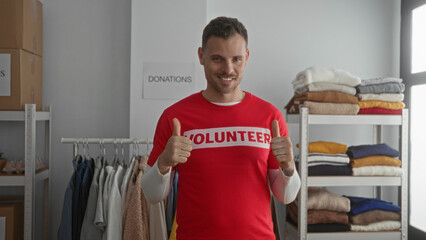 Young man in red volunteer shirt points and gives thumbs up indoors at a donation center surrounded by clothes and boxes, showing enthusiasm and dedication.