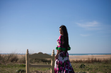 Latin woman brunette dancing flamenco in white dress with flower print and ruffles. The woman walks backwards and looks back at an old empty wooden chair. In the horizon the Atlantic Ocean.