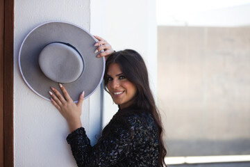 portrait of a Latin woman, young and beautiful flamenco dancer with her hands and arms on the wall holding a hat. The woman looks at the camera with a very happy face and smile.
