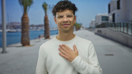 Man clutch chest in white sweater on street at seaside promenade lined with palm trees beside blue...
