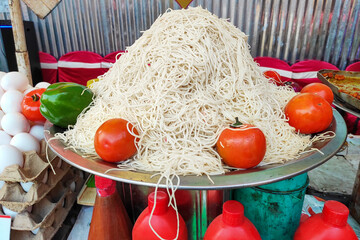 A vibrant street food stall features a heap of freshly boiled noodles on a steel platter, surrounded by red tomatoes. Trays of eggs and bottles of red sauce suggest delicious preparations ahead.