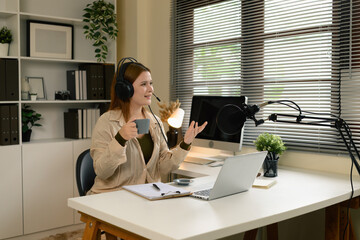 A Caucasian female podcaster hosting an online interview, wearing headphones and speaking into a microphone while enjoying a cup of coffee