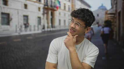 Young hispanic man wearing glasses hand on chin thinking on sunlit cobblestone street in historic...
