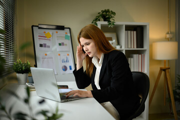 A Caucasian businesswoman looking stressed while working on a laptop in a modern office setting...