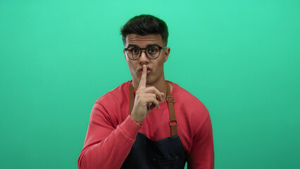Young man wearing glasses and a red apron gesturing silence against a green background with a...