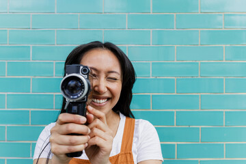 Smiling filmmaker holding vintage camera against turquoise brick wall