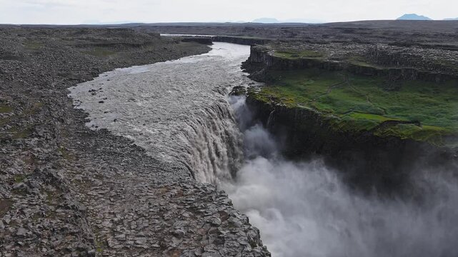 Dettifoss waterfall in Iceland shows intense water flow over rocky cliffs, creating mist. Rugged terrain and walking paths surround the area.