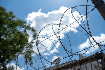 Barbed Wire Stretches Against a Bright Blue Sky With Clouds in a Secure Location Indicating...