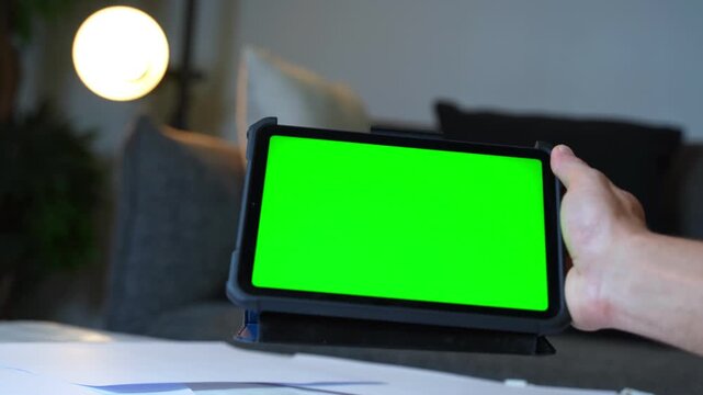 Hand holds tablet with green screen in warm home setting. Ideal for app previews, digital mockups, content creators, and tech promo scenes.