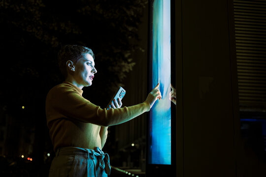 Woman using digital touch screen information display at night in city