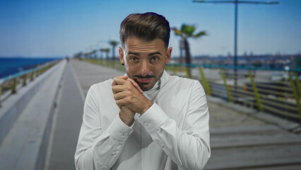 Man with white shirt and clasped hands on seaside promenade by palm trees and railing under clear...
