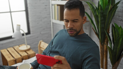 Fototapeta premium Young hispanic man in casual clothes relaxing on bed in cozy bedroom with smartphone and coffee enjoying leisure time at home indoors