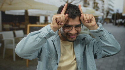 Young hispanic man in denim blue jacket and glasses finger pointing to head at sunlit restaurant...