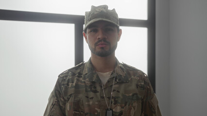 Hispanic military man in camouflage uniform with dogtags stands looking down by window in building...
