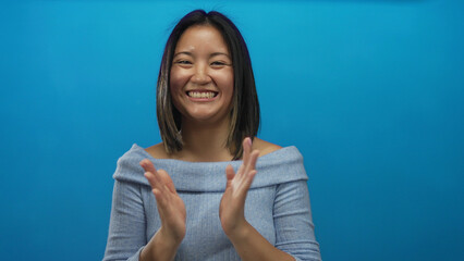 Fototapeta premium Woman clapping enthusiastically against an isolated blue background wall, showcasing joy and positivity with a vibrant smile, perfect for expressing happiness and success.
