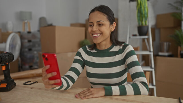 Woman smiling indoors while checking phone in new apartment surrounded by unpacked boxes ladder and tools representing a fresh start and joyful communication experience