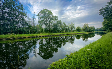 A beautifully serene view of a wide river with vibrant reflections and lush greenery under a cloudy sky above