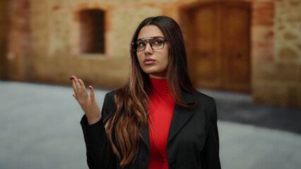 Fototapeta premium Young woman with glasses in red dress counts on fingers outdoors at an old university campus, signaling with focus and concentration.