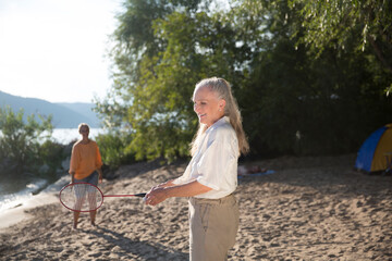 Summer lifestyle portrait senior woman with gray hair playing badminton, having fun in the seashore on sunny summer day.  Enjoying the little things. spends time in nature in summer.  women's friends