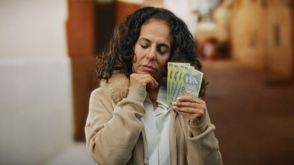 Middle-aged woman holding romanian lei banknotes, standing thoughtfully on a city street,...