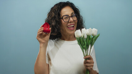 Hispanic woman holding bouquet and ring box against blue background, expressing joy for engagement...