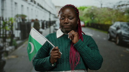 Fototapeta premium Woman outdoors holding a pakistan flag on a city street looking thoughtful, blending cultural diversity with an urban backdrop of cars and buildings.