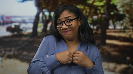 Woman in blue blouse and glasses shrugs with palms up in sunlit park lane under tall oak trees;...