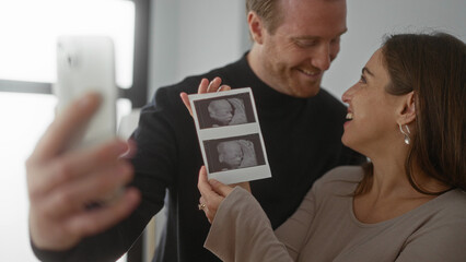 Couple joyfully taking selfie with ultrasound images in cozy home setting, highlighting excitement and anticipation of pregnancy journey together.
