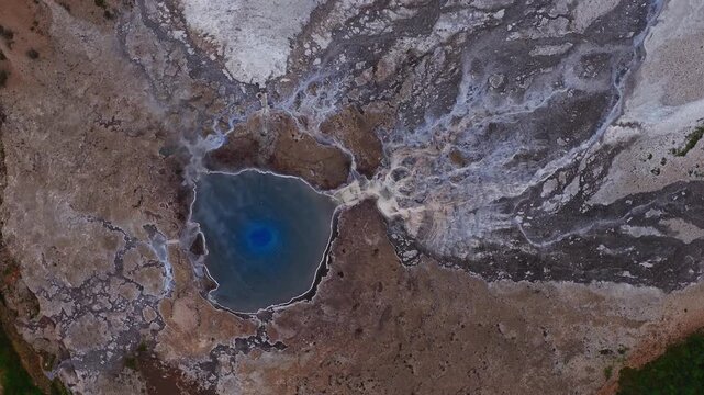 Aerial view of a vibrant blue geothermal hot spring in Iceland, surrounded by mineral deposits, rugged terrain, and patches of greenery. Steam rises visibly.
