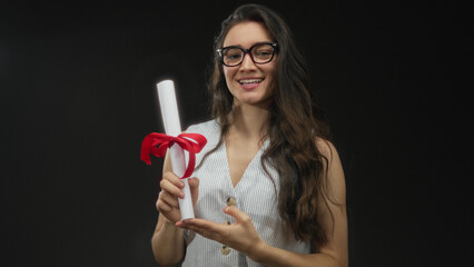 Young woman holding diploma scroll with red ribbon against a black studio backdrop; pride...