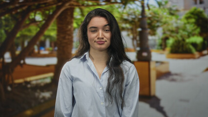 Young hispanic woman with long dark hair in striped shirt winks eye on leafy sunlit street corner;...