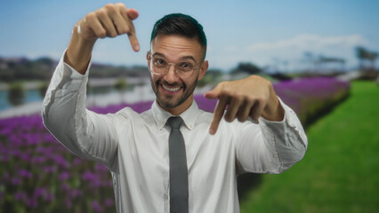 Smiling man in glasses and tie points downward in a vibrant green park with purple flowers under a blue sky, showcasing joyful outdoor business demeanor.
