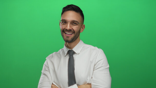 Young hispanic man smiling confidently with arms crossed against a vibrant green background wearing glasses and a formal white shirt paired with a stylish tie showcasing modern style.