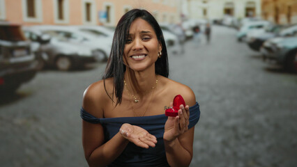 Woman outdoors holding engagement ring on a city street, expressing emotion, showing proposal...