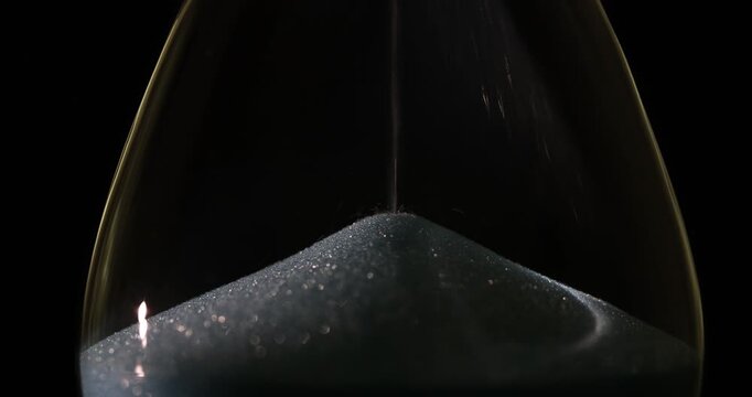 Sand trickles downward in constant stream marking passage of time. Grains of sand fall through narrow neck of hourglass and gather in lower chamber