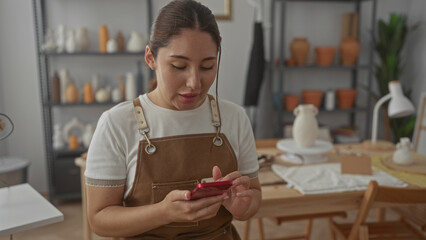 Young hispanic woman taps red smartphone in studio among clay pots and pottery tools; concentration...