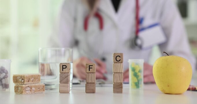 Miniature wooden cubes form abbreviation PFC on doctor table. Physician sits at desk with apple and writes prescription on sheet of paper. Slow motion