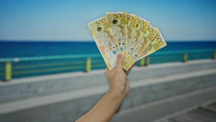 Man holding philippine pesos near seaside promenade with a clear blue sky in the background,...