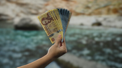Hand holding hungarian forint banknotes by the seaside, capturing a man's gesture outdoors against a beach background with scenic water view, implying financial travel themes. © Krakenimages.com