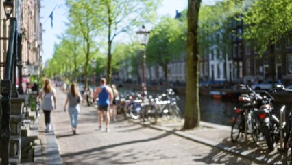 Blurred people walking alongside amsterdam canal with trees bicycles and historic buildings in the netherlands on a sunny day conveying a leisurely urban atmosphere.