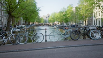 Fototapeta premium Blurred bicycles lined along an amsterdam canal with green trees and historic architecture in the background under a clear sky.