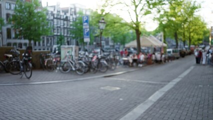 Blurred image of an amsterdam street scene showing people in the distance, with bicycles and historic buildings lining the cobblestone street in a vibrant city atmosphere.