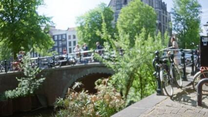 Blurred people walking along a picturesque amsterdam canal bridge with bicycles, trees, and historic architecture in a sunny, defocused scene.