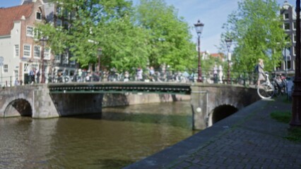Blurry view of a woman walking with a bicycle along a canal in amsterdam, netherlands, showcasing defocused urban scenery and iconic dutch architecture.