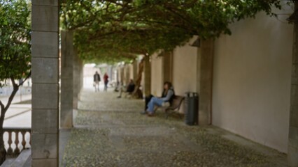 Blurred scene shows people relaxing on benches under shady trees in a university campus walkway, offering a serene and academic atmosphere with a touch of nature.