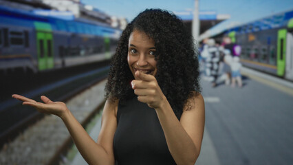 Smiling young hispanic woman pointing finger at train by building entrance; wanderlust excitement...