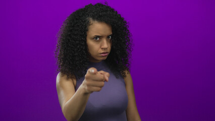 Young hispanic woman points finger at camera with furrowed brow against a vivid purple studio wall;...
