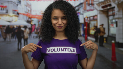 Latin woman points at purple volunteer shirt with thumbs up on busy city street during charity...
